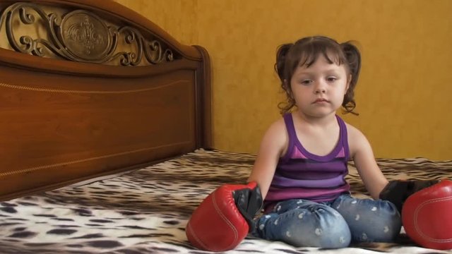A sad child in boxing gloves. A little girl is sitting on a bed in gloves for boxing.