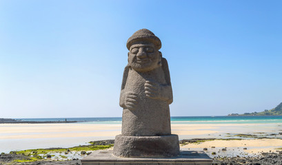 statue of Dol hareubang - symbol of Jeju island on sunny day with sea and beach on background