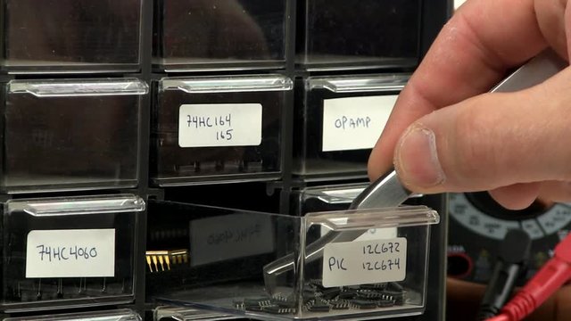 Hand opens circuit component supply draw bin to get surface mount micro-controllers.  Components grabbed with tweezers.  Draw is then closed.  DVM voltage/current meter is visible in background.