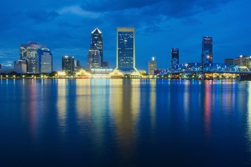 Fototapeta premium View of a downtown at dusk with buildings, bridge and colored lights reflecting over water shot as long exposure from across the water with blue sky, landscape composition