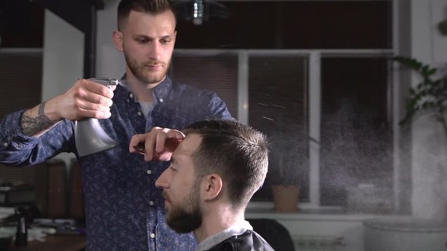 The hairdresser splashes water from the atomizer on the head of the visitor to the barbershop who is waiting for the haircut to end