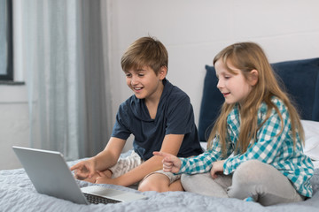 Small Boy And Girl Use Laptop Computer Sitting On Bed In Bedroom, Brother And Sister Surfing Internet Together