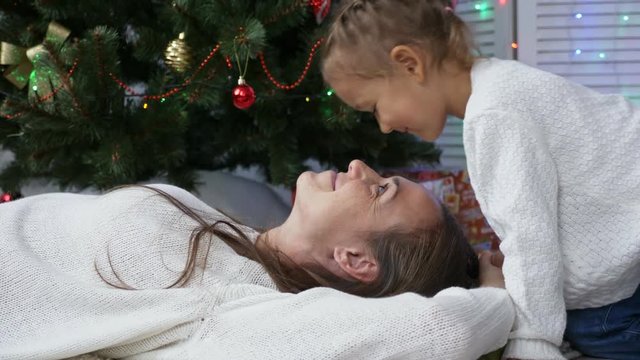Cute little girl having fun and kissing her mom next to Christmas tree