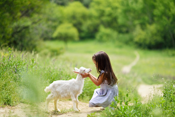 Little girl plays and huhs goatling in country, spring or summer nature outdoor. Cute kid with baby animal, forest, trod, glade background. Friendship of child and yeanling, image toned.