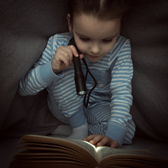 Little girl reading fairy tales book under the covers at the evening with lantern. Cute kid playing...