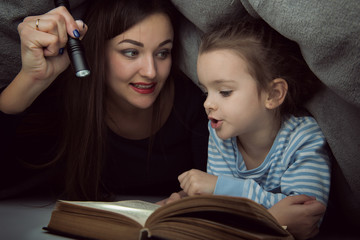 Little girl and her mother reading fairy tales book under the covers at the evening with lantern. Cute kid playing before going to sleep, image toned. Home family leisure.