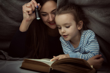 Little girl and her mother reading fairy tales book under the covers at the evening with lantern....