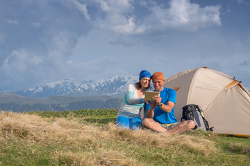 Naklejka na ściany i meble Joyful travelers sit next to tent use tablet and having fun