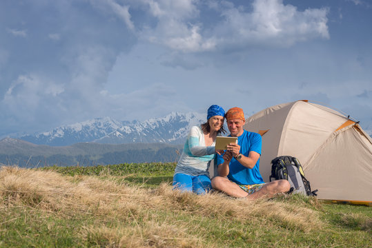 Joyful travelers sit next to tent use tablet and having fun