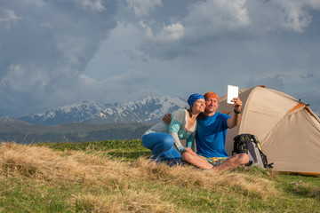 Fototapeta na wymiar Funny travelers sit next to tent, use table, taking selfie