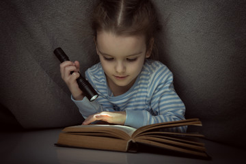 Little girl reading fairy tales book under the covers at the evening with lantern. Cute kid playing...