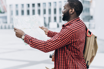 Handsome and happy Afro American tourist with old camera and map in modern city looking at the map