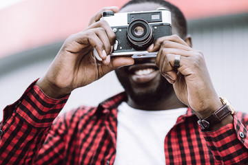Outdoor summer smiling lifestyle portrait of handsome and happy Afro American tourist having fun in the city in Europe with camera travel photo of photographer making pictures in hipster style