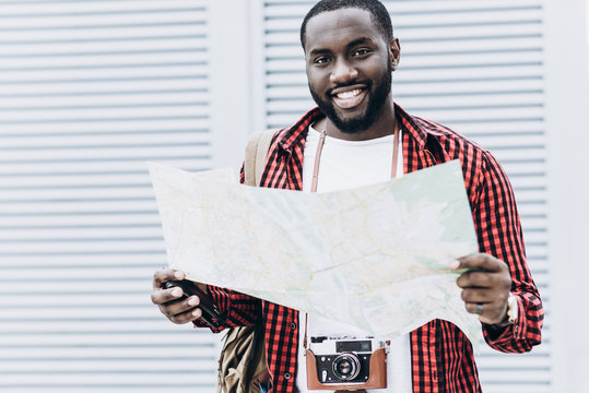 Portrait of handsome Afro American tourist wearing casual clothes with old camera and map in modern city. He is looking at camera and smiling