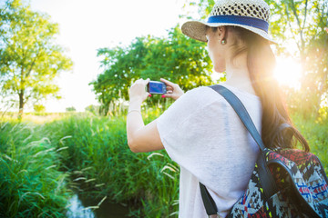 Beautiful caucasian model standing in the nature on a footbridge over small stream wearing casual summer clothes, straw hut and backpack, taking photo with a small digital camera. Summer sunset.