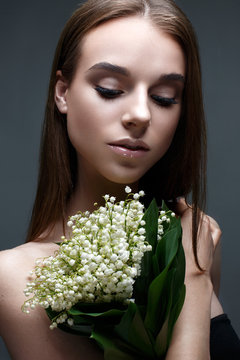A young girl with a bouquet of lilies of the valley and straight flowing hair. A beautiful model with a gentle nude make-up and shining skin. Photo is taken in the studio. Beauty of the face.