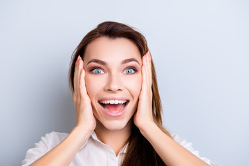 Close up cropped portrait of cute amazed girl in white shirt, holding her head, she is shocked, extremely happy, with wide open eyes and mouth on pure grey background