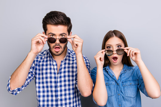 Oh really?! Wow! Two young shocked lovers are amazed with wide open eyes, mouthes, touching sun glasses, standing in a casual wear on light background