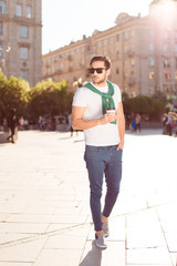 Fototapeta na wymiar Great weekend. Full length of harsh young brunete guy, having hot tea and walking in the city outdoors, in casual comfortable outfit, trendy sunglasses