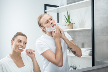 portrait of wife brushing teeth and looking at husband putting foam on face