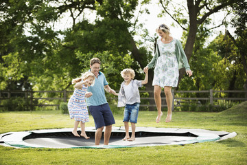 Man, woman, boy and girl holding hands, jumping on a trampoline set into the ground in a garden.