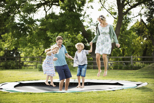 Man, woman, boy and girl holding hands, jumping on a trampoline set into the ground in a garden.