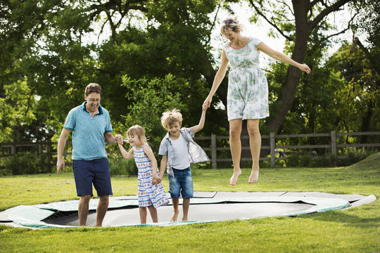 Man, woman, boy and girl holding hands, jumping on a trampoline set in the lawn in a garden.