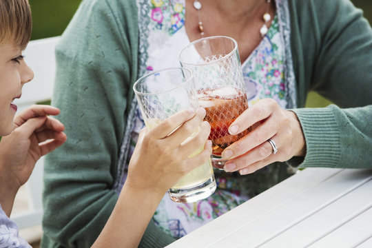 Woman And Boy Sitting Outdoors At A Table, Holding Drinks Glasses, Clinking Them Together. 