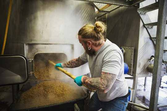 Man scraping spent grain from a large kettle in a brewery.