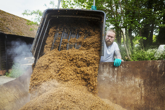 Man pouring spent grain into a large container, steam rising, during the brewing process. 