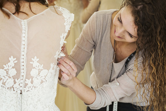 A young woman in a white wedding dress, and a dressmaker fitting the bodice and waist, adjusting the sizing. 