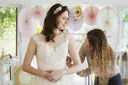 A young woman in a white wedding dress, and a dressmaker fitting the bodice and waist, adjusting the sizing. 