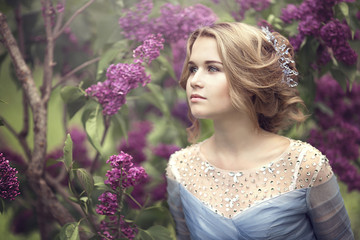 Portrait of a beautiful young blond woman in lilac bushes, admiring flowers.