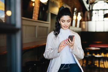 Pretty brunette woman with pony tail and appealing appearance sitting in cafe using her smartphone surfing internet using free wifi conncection and listening to her favourite music with earphones