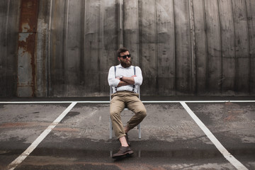 fashionable handsome man in sunglasses and suspenders sitting on parking place