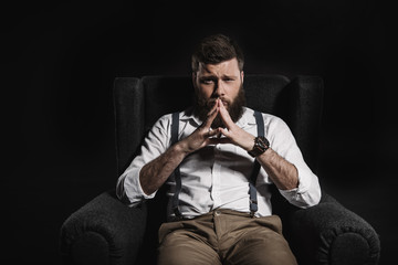 thoughtful fashionable businessman sitting in armchair and looking at camera, isolated on black