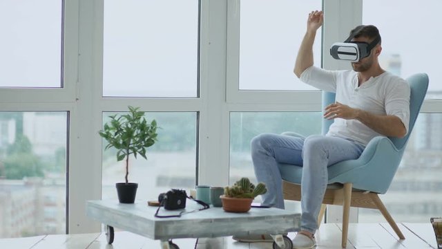 Young man have VR experience using virtual reality headset sitting in chair on balcony in loft apartment