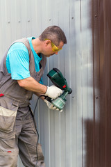 Man in overalls and protective glasses with spray gun in hand. Painting of metal fence