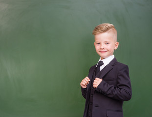 Happy boy in a suit standing near empty green chalkboard