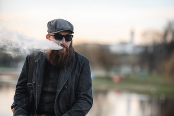 Vape bearded man in real life. Portrait of young guy with large beard in a cap and sunglasses vaping an electronic cigarette with puffs of steam on the embankment near the river in the spring evening.