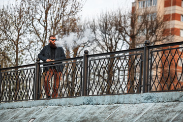 Vape bearded man in real life. Portrait of young guy with large beard in sunglasses vaping an electronic cigarette with puffs of steam on the embankment at sunset in the summer. Lifestyle.