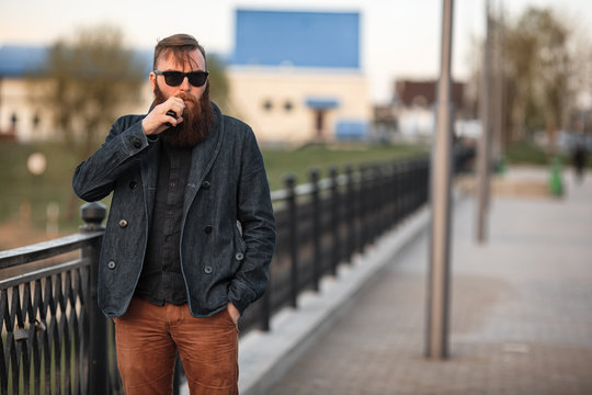 Vape bearded man in real life. Portrait of young guy with large beard in sunglasses vaping an electronic cigarette on the embankment near the river in the spring evening.