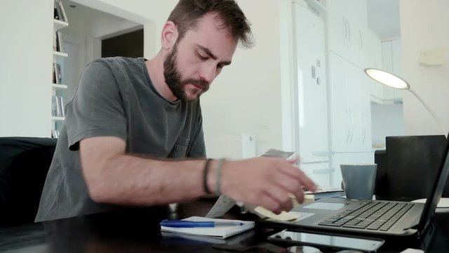 A young man at home calculates his living expenses and sums his bills and his overdue notices and falls into despair