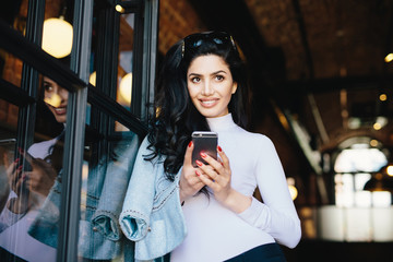 Attractive woman with dark hair, bright eyes and long eyelashes having sunglasses on head looking pensively up having some dreams while communicating with her lover via social networks, using Internet