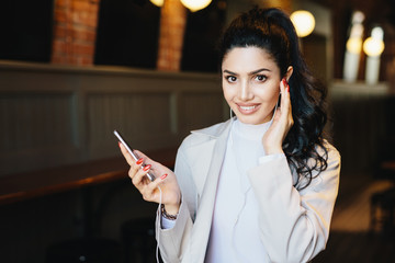 Cute woman with dark pony tail having long eyelashes and expressive eyes sitting at cafe listening to music online enjoying high-speed internet connection. People, beauty and technology concept