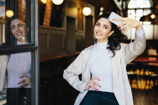 Waist up portrait of beautiful lady with dark hair tied in pony tail wearing white jacket sitting in cafe making selfie using her modern smartphone and listening to music with earphones. People, rest