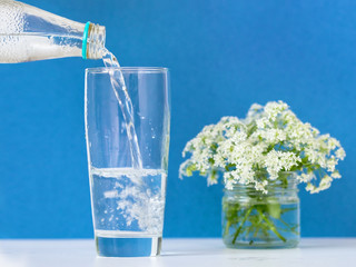 Pure drinking water poured from a bottle into a glass Cup on the table. Blue background. Vase with...