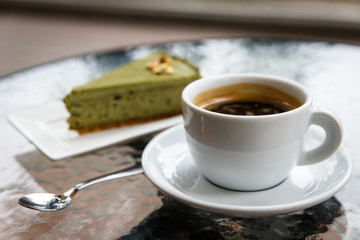 A pistachio cheesecake on a plate, a white cup of black coffee, a tea spoon. On a glass table top. Selective focus.