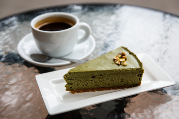 A pistachio cheesecake on a plate, a white cup of black coffee, a tea spoon. On a glass table top. Selective focus.