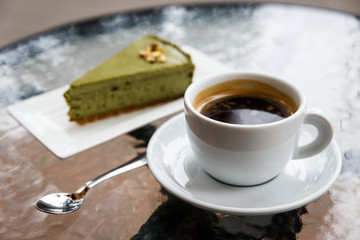 A pistachio cheesecake on a plate, a white cup of black coffee, a tea spoon. On a glass table top. Selective focus.